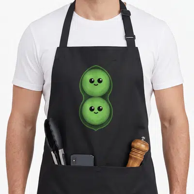 Peas pod aprons - Two cartoon peas with smiling faces stacked vertically inside a green pod — shown on a black cotton canvas apron against a white background