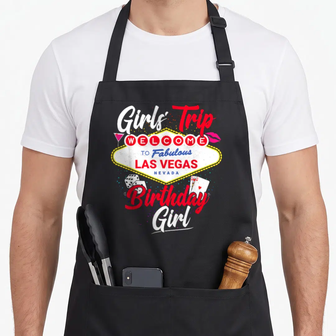 Souvenir birthday apron - Las Vegas sign graphic with Girls Trip text, cocktail glass, lips, dice, and playing cards — shown on a black cotton canvas apron against a white background
