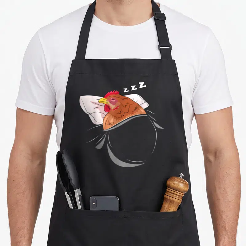 Sleeping chicken apron - Cartoon brown chicken lying on white pillow with ZZZ sleep text floating above head — shown on a black cotton canvas apron against a white background