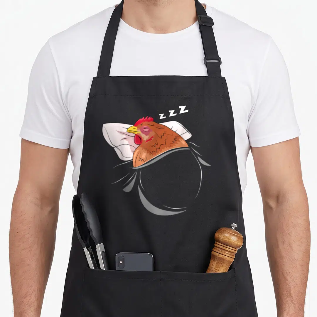 Sleeping chicken apron - Cartoon brown chicken lying on white pillow with ZZZ sleep text floating above head — shown on a black cotton canvas apron against a white background