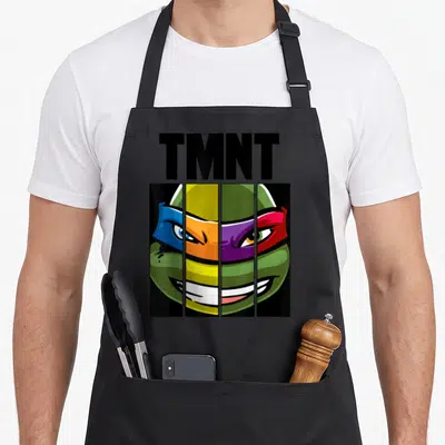 Mash face apron - TMNT text above turtle face split into four vertical panels with blue orange purple red green — shown on a black cotton canvas apron against a white background
