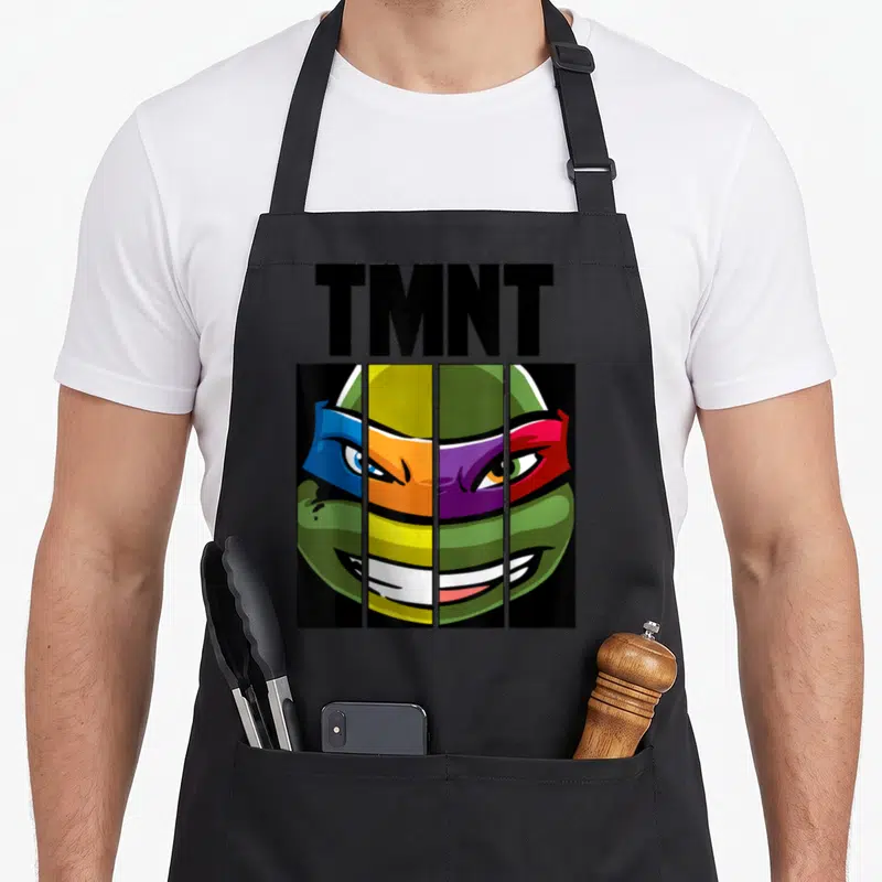 Mash face apron - TMNT text above turtle face split into four vertical panels with blue orange purple red green — shown on a black cotton canvas apron against a white background