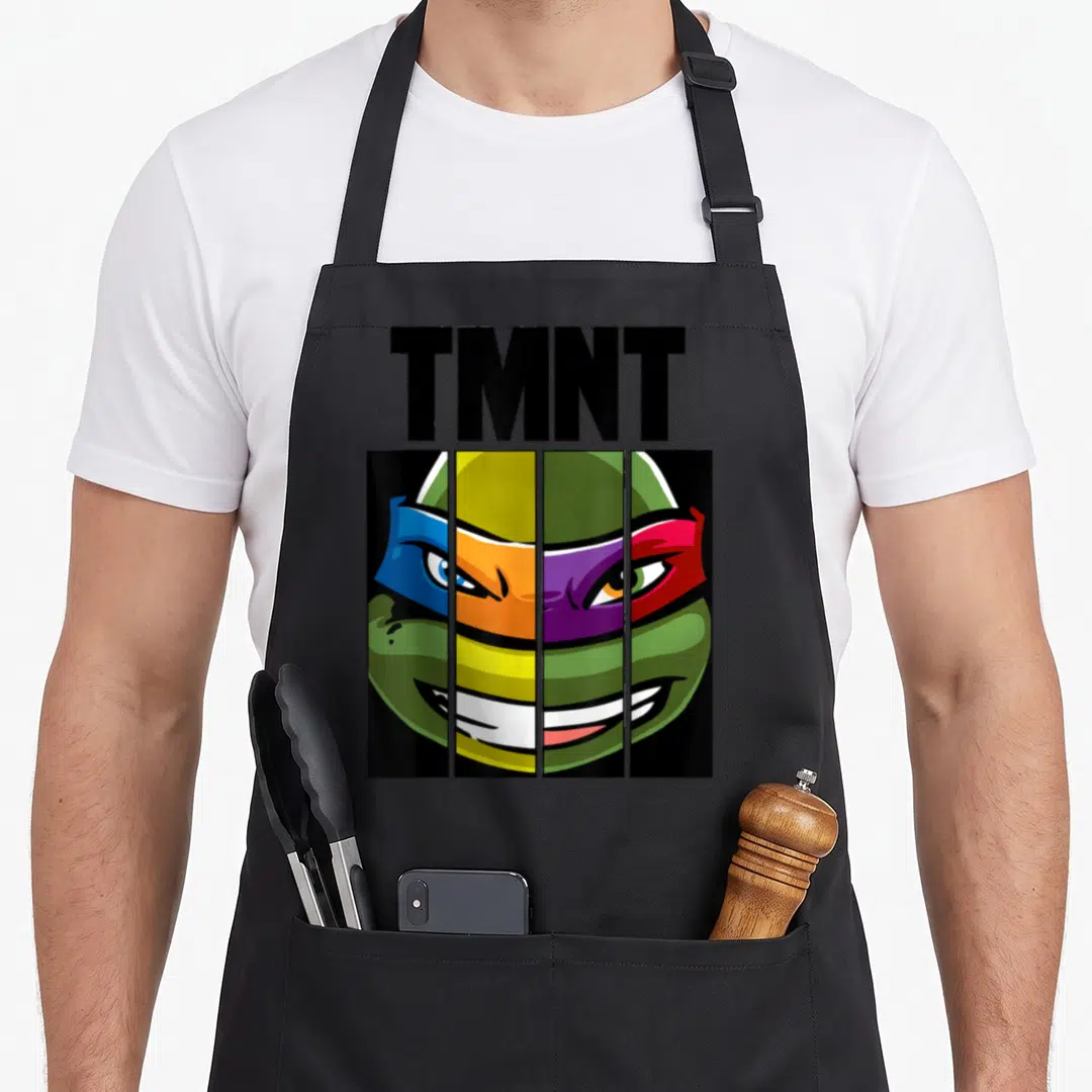 Mash face apron - TMNT text above turtle face split into four vertical panels with blue orange purple red green — shown on a black cotton canvas apron against a white background