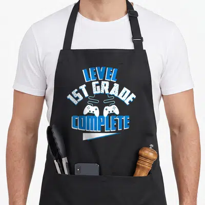 Grade level apron - LEVEL 1ST GRADE COMPLETE text with two white game controllers and blue banner — shown on a black cotton canvas apron against a white background