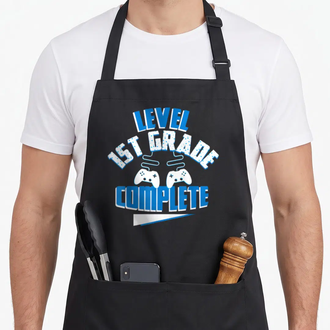 Grade level apron - LEVEL 1ST GRADE COMPLETE text with two white game controllers and blue banner — shown on a black cotton canvas apron against a white background