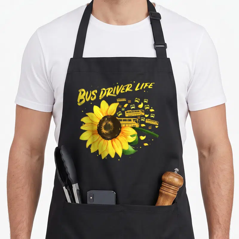 Bus sunflower apron - Yellow sunflower with small buses and arched text in watercolor style — shown on a black cotton canvas apron against a white background