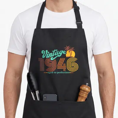 Cute 75th birthday apron - Retro 1946 text with palm trees and sun in teal, red, orange, and yellow — shown on a black cotton canvas apron against a white background