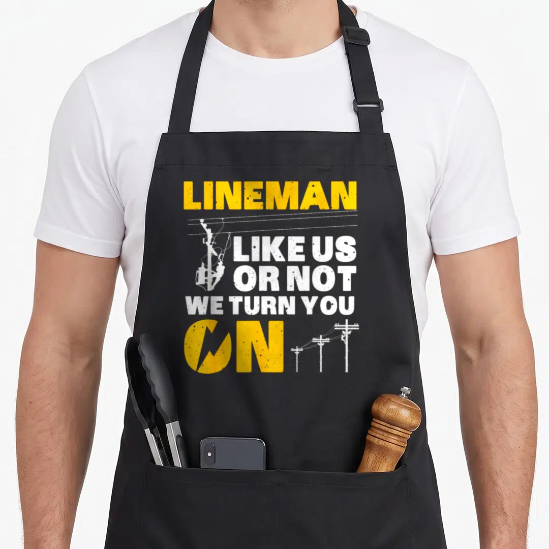 Electronic electricians apron - Yellow and white text reading Lineman Like Us Or Not We Turn You On with utility pole — shown on a black cotton canvas apron against a white background