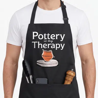 Pottery my therapy apron - Stacked white text reading Pottery is my Therapy above a cartoon terracotta pot on a wheel — shown on a black cotton canvas apron against a white background