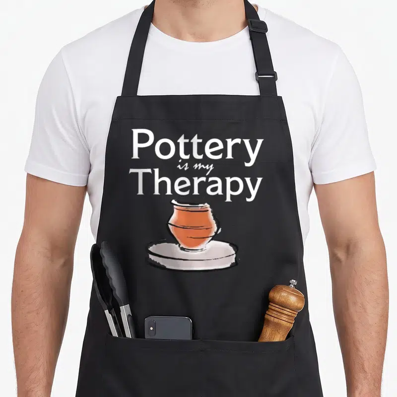 Pottery my therapy apron - Stacked white text reading Pottery is my Therapy above a cartoon terracotta pot on a wheel — shown on a black cotton canvas apron against a white background