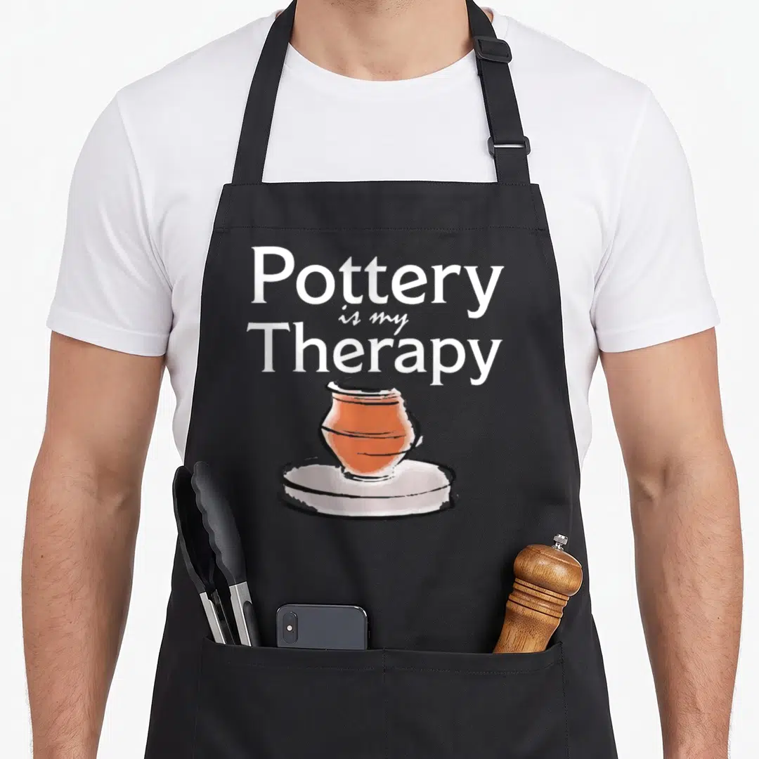 Pottery my therapy apron - Stacked white text reading Pottery is my Therapy above a cartoon terracotta pot on a wheel — shown on a black cotton canvas apron against a white background