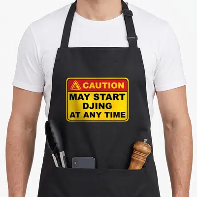 Caution dj quotes apron - Yellow caution sign with red header and black text reading May Start DJing At Any Time — shown on a black cotton canvas apron against a white background