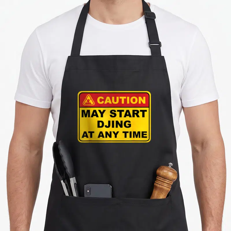 Caution dj quotes apron - Yellow caution sign with red header and black text reading May Start DJing At Any Time — shown on a black cotton canvas apron against a white background