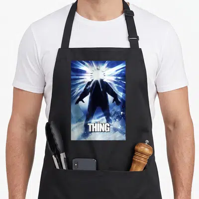Glowing movie poster apron - Silhouette figure with white light burst from head and THE THING text at bottom — shown on a black cotton canvas apron against a white background