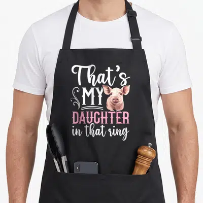 Daughter cooking mama apron - Cartoon pig head next to stacked white and pink text reading That's MY DAUGHTER in that — shown on a black cotton canvas apron against a white background