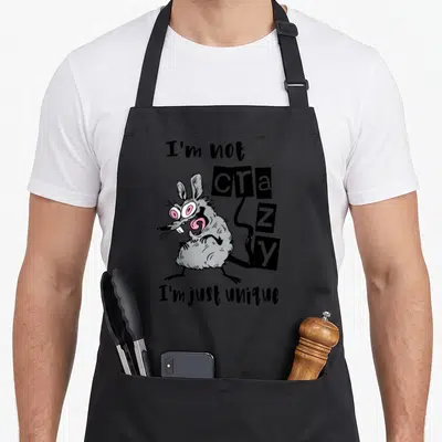 Mouse unique apron - Cartoon grey rat with pink eyes and text I'm not crazy I'm just unique — shown on a black cotton canvas apron against a white background