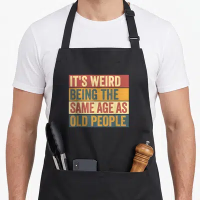 Age odd aprons - Retro red orange blue beige stripes with stacked text IT'S WEIRD BEING THE SAME AGE AS OLD PEOPLE — shown on a black cotton canvas apron against a white background