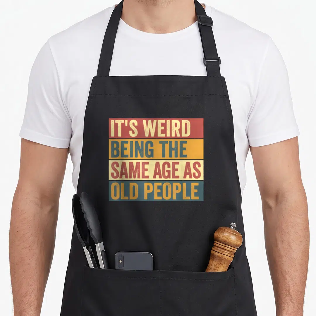 Age odd aprons - Retro red orange blue beige stripes with stacked text IT'S WEIRD BEING THE SAME AGE AS OLD PEOPLE — shown on a black cotton canvas apron against a white background