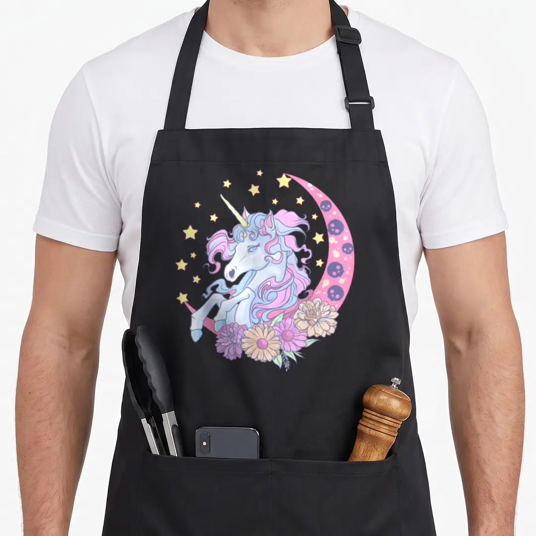 Unicorn anime cooking apron - Cartoon unicorn with pink mane, purple skulls in pink moon, yellow stars, and flower — shown on a black cotton canvas apron against a white background