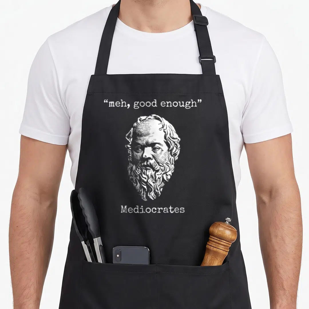 Mediocrate meme apron - White line art of bearded man head between meh good enough and Mediocrates text — shown on a black cotton canvas apron against a white background