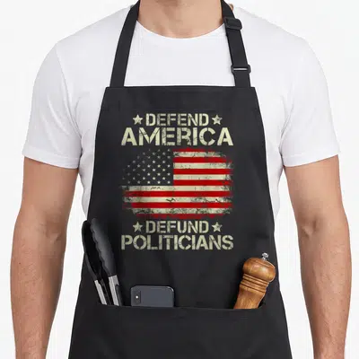Flag politician apron - Distressed USA flag between Defend America and Defund Politicians stencil text — shown on a black cotton canvas apron against a white background
