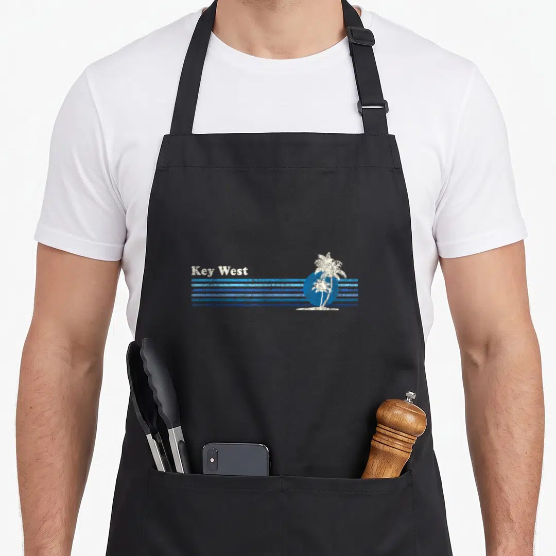 Key tree apron - Key West text white palm tree blue sun distressed stripes — shown on a black cotton canvas apron against a white background