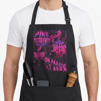 Parade elephant print apron - Pink elephants on parade text with cartoon characters, bubbles, and wavy lines in pink — shown on a black cotton canvas apron against a white background