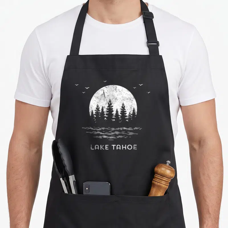 Tahoe tree apron - Distressed white sun behind pine trees and water lines above LAKE TAHOE text — shown on a black cotton canvas apron against a white background