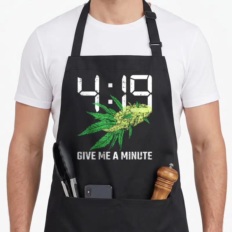 Give hemp apron - Distressed white 4:19 text over green cannabis bud with GIVE ME A MINUTE caption — shown on a black cotton canvas apron against a white background
