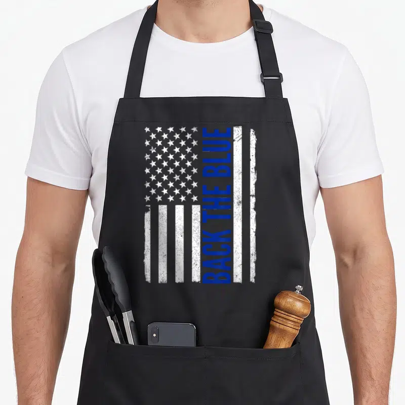 Blue pro apron - Vertical Back The Blue text on distressed white and blue flag with stars — shown on a black cotton canvas apron against a white background