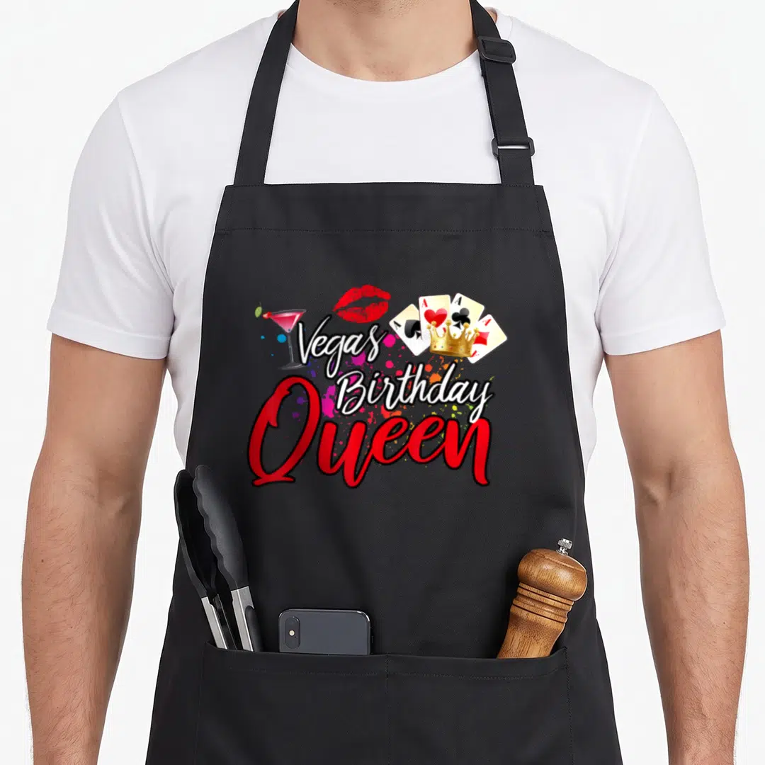 Vegas friends apron - Vegas Birthday Queen text with cocktail, lips, crowning playing cards, and paint splatters — shown on a black cotton canvas apron against a white background