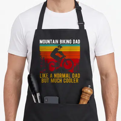 Mountain like normal apron - Retro biker silhouette on striped sunset with MOUNTAIN BIKING DAD LIKE A NORMAL DAD — shown on a black cotton canvas apron against a white background