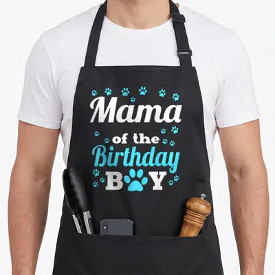 Birthday dog apron - Mama of the Birthday BOY text with blue paw print replacing the letter O — shown on a black cotton canvas apron against a white background