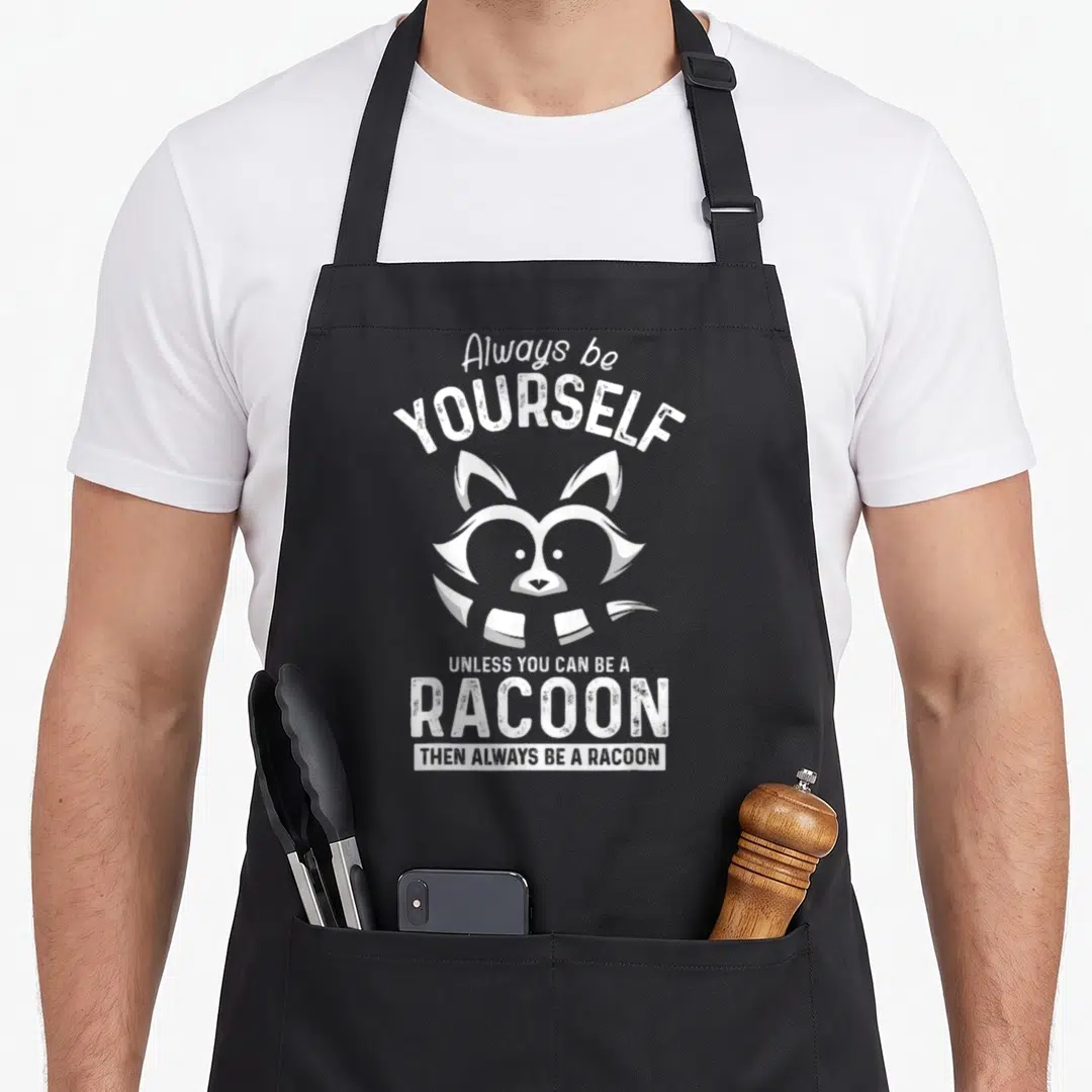 Sei raccoon apron - White distressed raccoon head with arched 'Always be yourself unless you can be a racoon' text — shown on a black cotton canvas apron against a white background