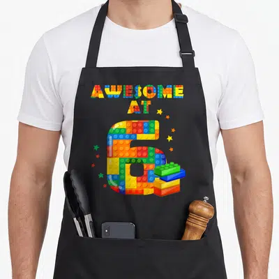 Birthday building block & apron - AWESOME AT 6 brick text with stacked blocks and scattered stars in primary colors — shown on a black cotton canvas apron against a white background