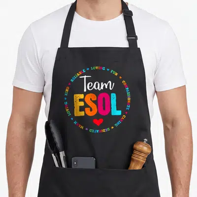Crew cute teacher aprons - Team ESOL text surrounded by loving fun compassionate caring dedicated warm kind reliable — shown on a black cotton canvas apron against a white background