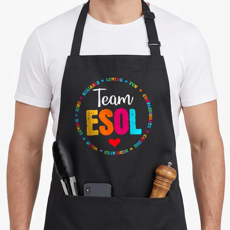 Crew cute teacher aprons - Team ESOL text surrounded by loving fun compassionate caring dedicated warm kind reliable — shown on a black cotton canvas apron against a white background