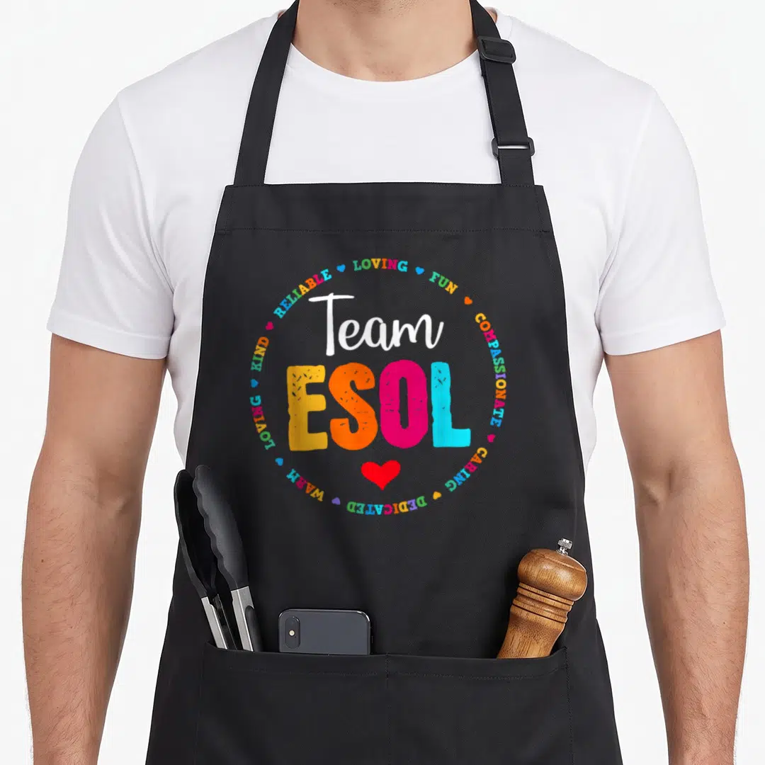 Crew cute teacher aprons - Team ESOL text surrounded by loving fun compassionate caring dedicated warm kind reliable — shown on a black cotton canvas apron against a white background