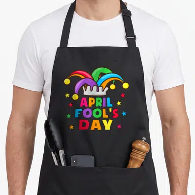 April prank apron - Cartoon jester hat with red blue yellow green purple colors above stacked text and scattered stars — shown on a black cotton canvas apron against a white background