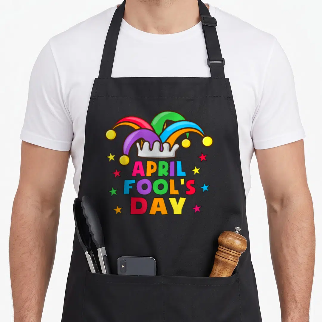 April prank apron - Cartoon jester hat with red blue yellow green purple colors above stacked text and scattered stars — shown on a black cotton canvas apron against a white background