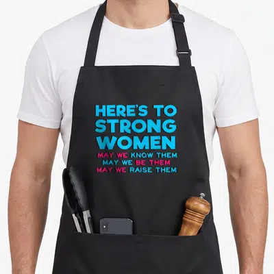 Motivational strong mom apron - Stacked blue and pink text reading HERE'S TO STRONG WOMEN with three supporting lines — shown on a black cotton canvas apron against a white background