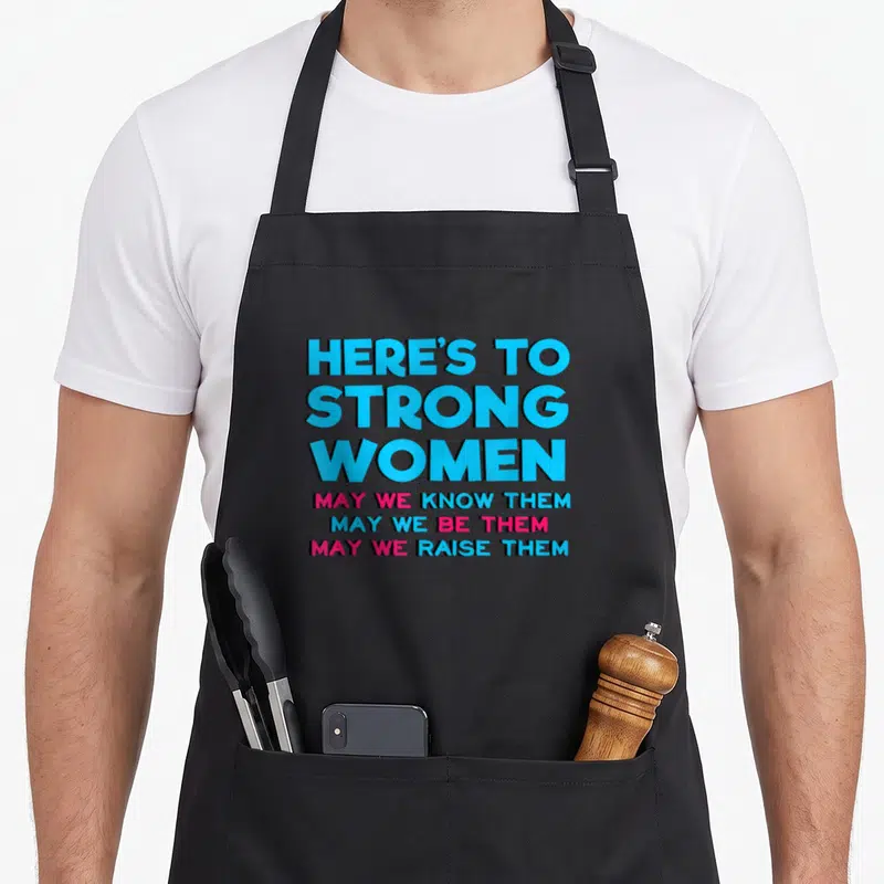 Motivational strong mom apron - Stacked blue and pink text reading HERE'S TO STRONG WOMEN with three supporting lines — shown on a black cotton canvas apron against a white background