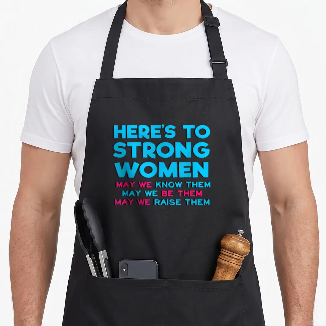 Motivational strong mom apron - Stacked blue and pink text reading HERE'S TO STRONG WOMEN with three supporting lines — shown on a black cotton canvas apron against a white background