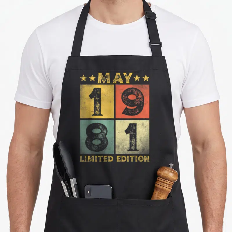 May bday birthday apron - MAY 1981 LIMITED EDITION text in yellow red green blue-grey retro grid — shown on a black cotton canvas apron against a white background
