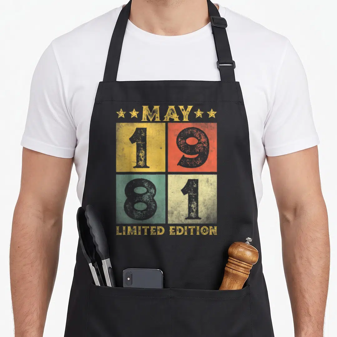 May bday birthday apron - MAY 1981 LIMITED EDITION text in yellow red green blue-grey retro grid — shown on a black cotton canvas apron against a white background
