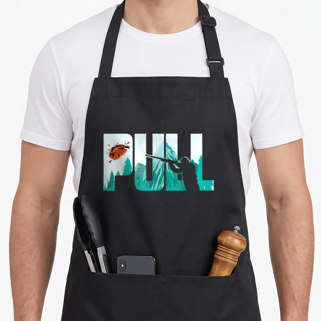 Pull clay apron - PULL text containing teal mountains, hunter silhouette, and orange breaking clay target — shown on a black cotton canvas apron against a white background