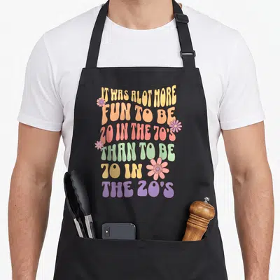 Retirement 70th birthday apron - Retro wavy text reading 20 in 70s 70 in 20s with four pink flowers — shown on a black cotton canvas apron against a white background
