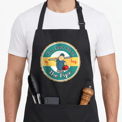 Plumber lay pipe apron - Cartoon plumber holding pipe and toolbox inside teal and yellow circle with arched text — shown on a black cotton canvas apron against a white background