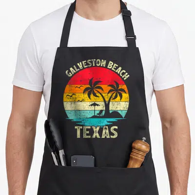 Family vacation texas apron - Retro Galveston Beach Texas sunset with palm tree, beach chair, umbrella, and seagull — shown on a black cotton canvas apron against a white background