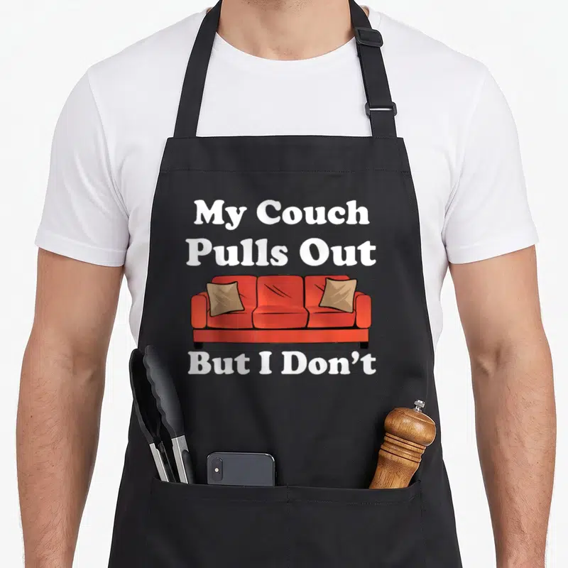 Pull couch apron - Red cartoon couch with two tan pillows and white text reading My Couch Pulls Out But I Don't — shown on a black cotton canvas apron against a white background
